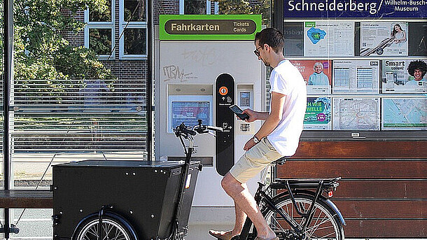 Ein Lastenrad-Fahrer steht an einer Stadtbahn-Haltestelle vor einem Fahrkartenautomaten.